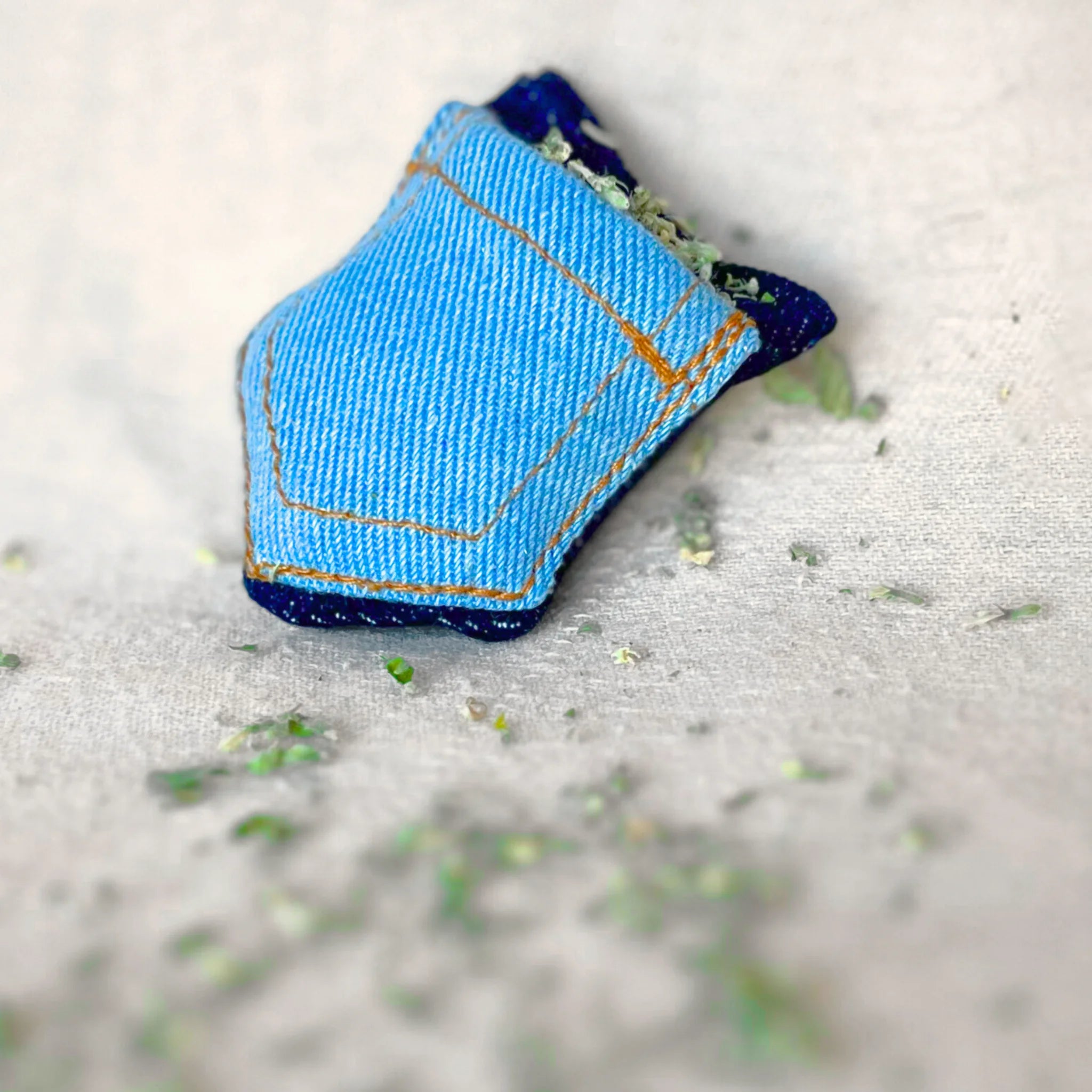 Close-up of a small denim pocket cat toy lying on a surface with loose catnip scattered around.