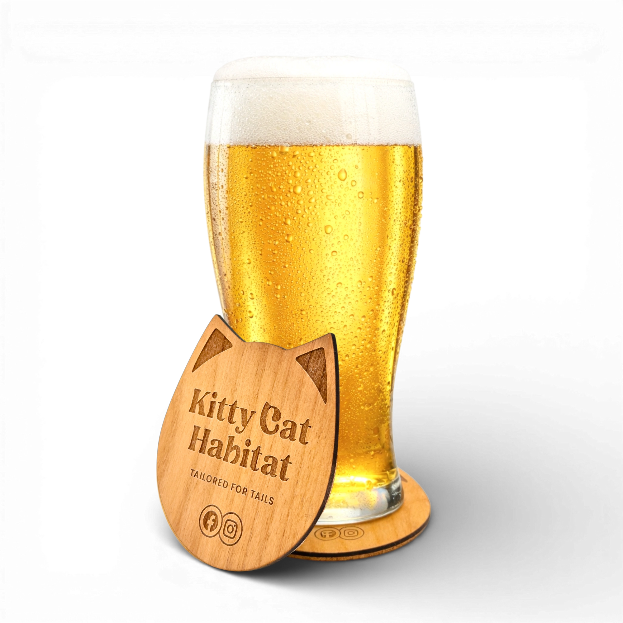 Beer glass resting on a wooden cat head drink coaster with a second Kitty Cat Habitat logo coaster in front on a light background.